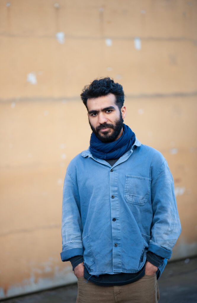 Porträt von Eslam Elnebishy. Er ist ein junger Mann mit dunklen Haaren und einen Bart. Er trägt ein blaues Hemd und einen blauen Schal.