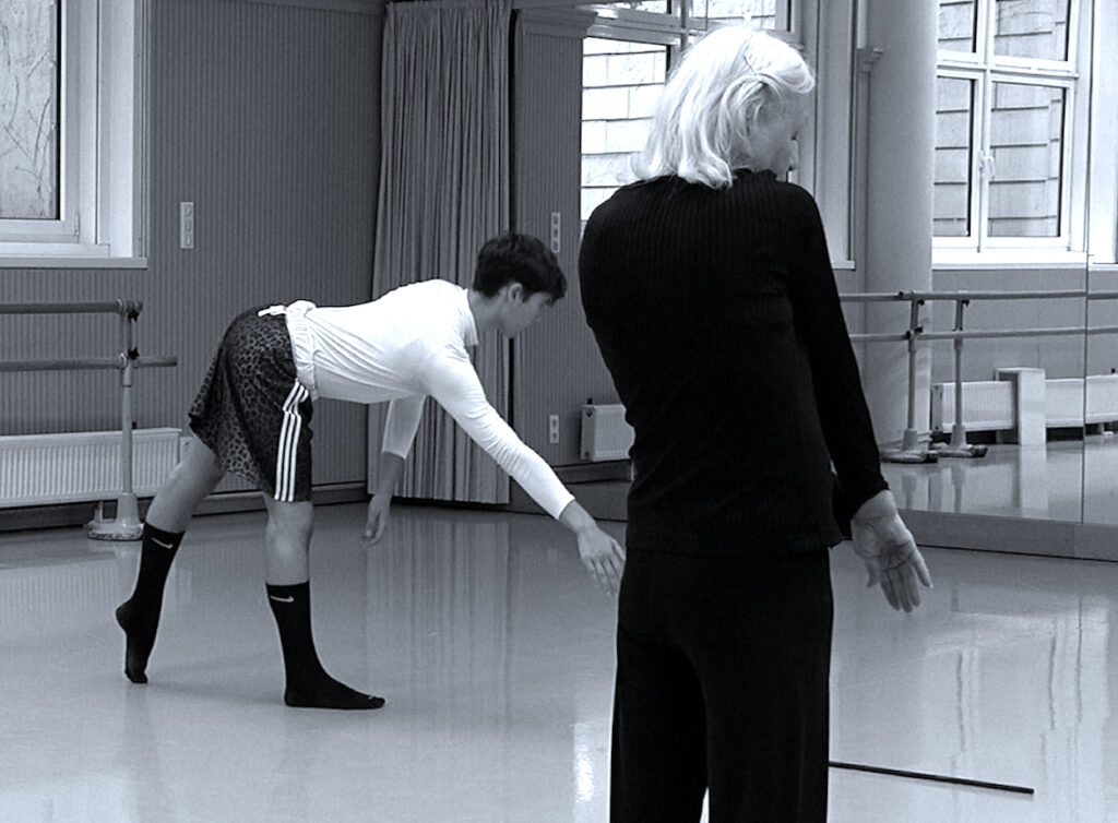 Das Bild ist Schwarz-Weiß. Im Vordergrund steht eine alte Frau mit weißen Haaren und schwarzer Kleidung mit dem Rücken zur Kamera. Im Hintergrund auf der linken Seite des Bildes, tanzt eine junge Person mit kurzen Haaren. Sie sind in einem Tanzstudio vor einer Spiegelwand.