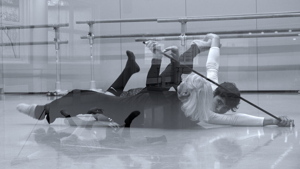 Bild in schwarzweiß. Eine ältere Frau mit schwarzer Kleidung und ein junger Mann mit weißer Kleidung bewegen sich zusammen im Raum. Das Bild ist verspiegelt.