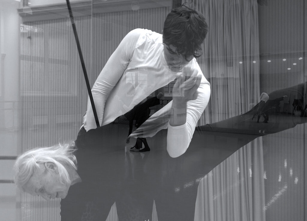 Bild in schwarzweiß. Eine ältere Frau mit schwarzer Kleidung und ein junger Mann mit weißer Kleidung bewegen sich zusammen im Raum. Das Bild ist verspiegelt.