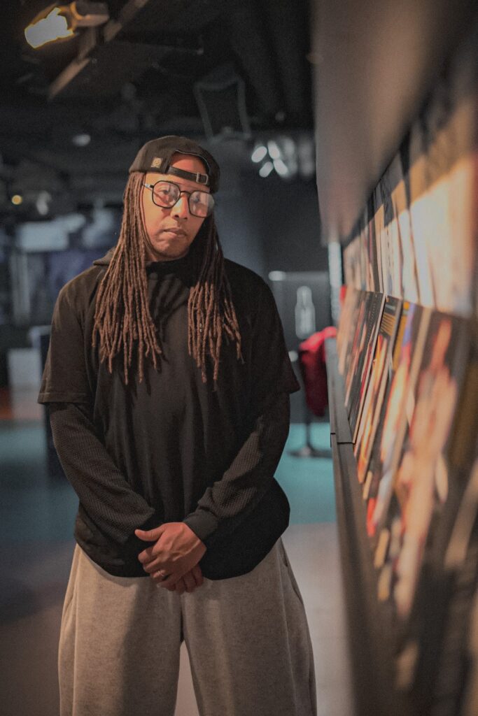 Born von HOUSEaHOLICS posiert neben einem Plattenregal. Er trägt eine schwarze Cap rückwärts, eine Brille und einen schwarzen Hoodie. ER hat Dreads die bis zur Brust reichen. Seine Hände liegen locker vor dem Körper zusammen.