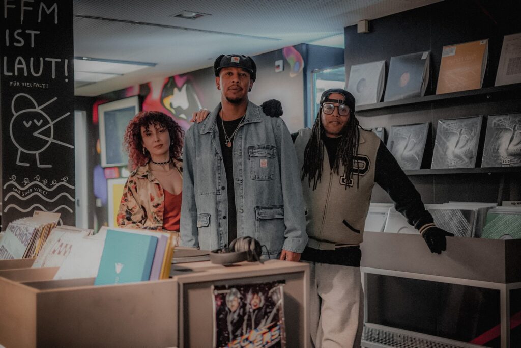 Die drei Mitglieder von HOUSEaHOLICS stehen vir einem Plattenregal und schauen in die Kamera.