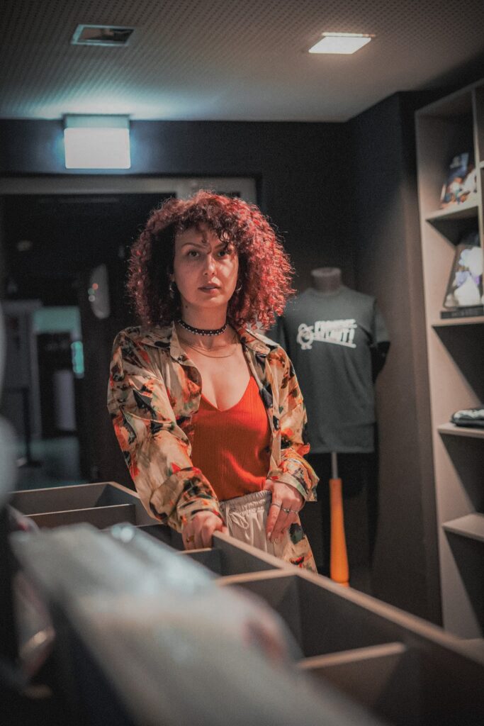 Nora von HOUSEaHOLICS steht neben einem Plattenregal und guckt in die KAmera. Sie braun-rötliche Locken, trägt ein oragendes Top und darüber eine bunte Bluse.