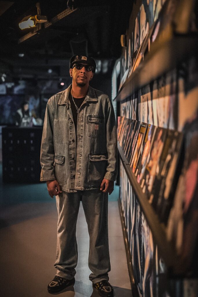 Kevin von HOUSEaHOLICS steht neben einem Plattenregal und guckt in die Kamera. Er trägt eine schwarze Cap und eine Sonnenbrille. Seine Haare sind kurz. Er trägt eine Jeans-Jacke und eine Jeans-Hose.