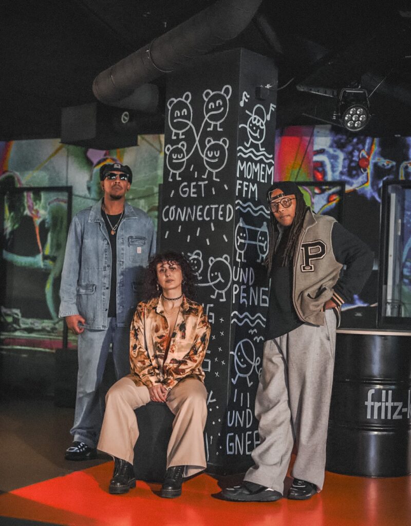 Auf dem Bild sind drei Personen zu sehen, die sich in einem bunten Raum befinden. In der Mitte befindet sich eine schwarze Säule, auf die mit weißer Schrift kleine Zeichnungen und Sätze gemalt sind. Einer der Sätze ist: Get connected. Die linke Person steht neben der Säule und trägt Jacke und Hose aus Jeansstoff. Die Person in der Mitte sitzt vor der Säule auf einem Hocker. Sie trägt eine lockere beige Hose und ein buntes Hemd. Die Rechte Person ist an die Säule gelehnt und trägt eine lockere Graue Jogginghose, eine schwarze Kappe, eine Brille, einen schwarzen Pullover und eine Weste darüber.