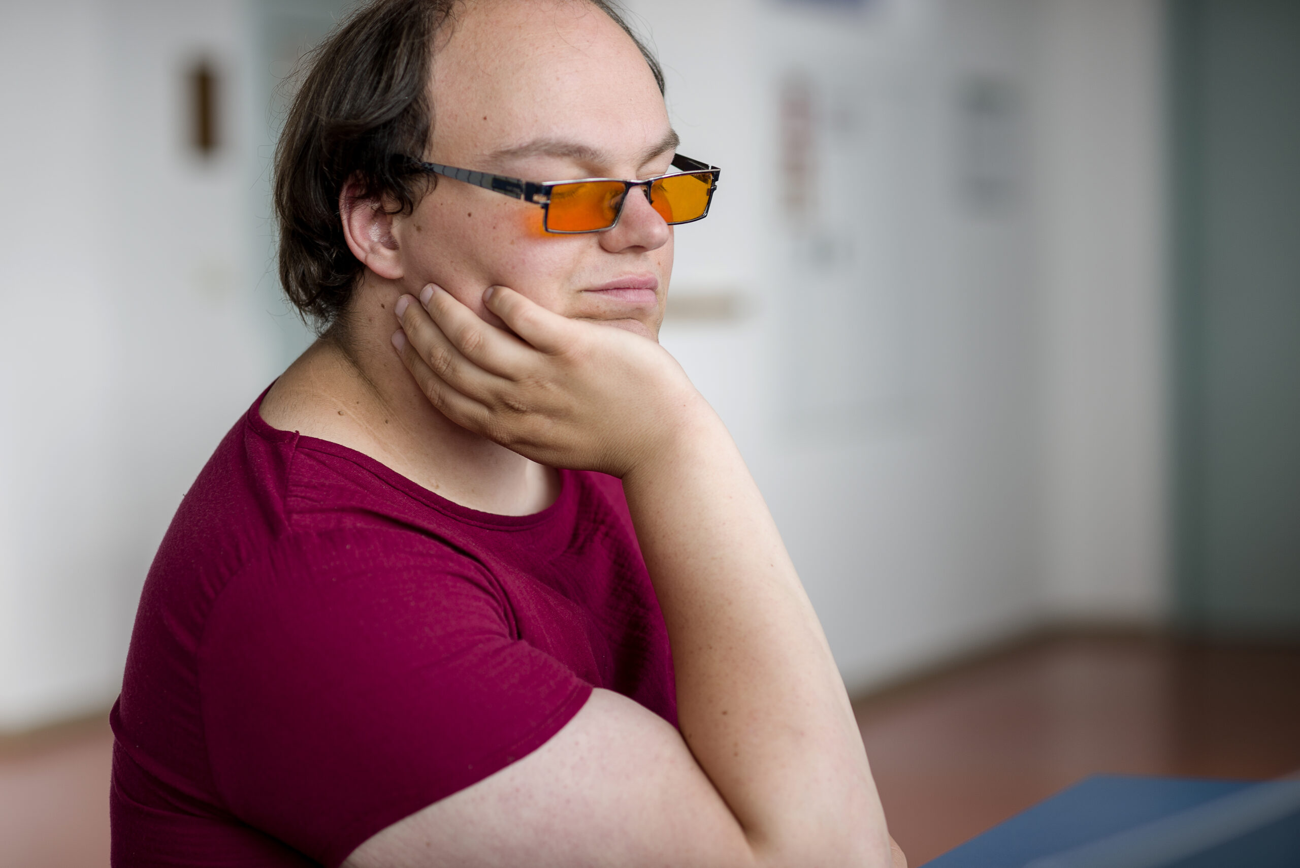 Eine Person hält den Kopf auf die Hand gestützt. Sie trägt ein rotes T-Shirt und eine Brille mit orangenen Gläsern.