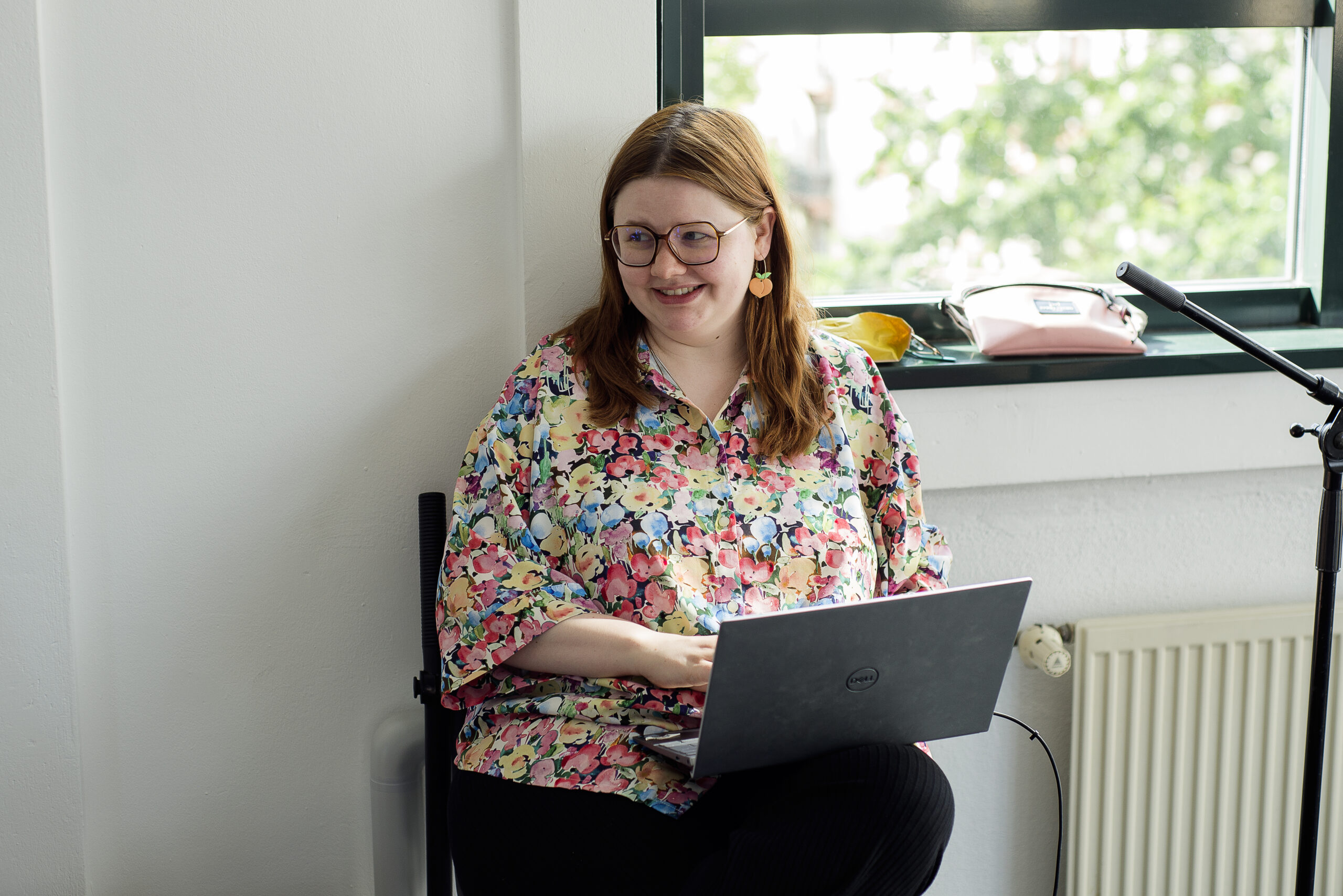 Eine Person sitzt lächelnd auf einem Stuhl neben einem Fenster. Sie hat rote Haare, trägt eine Brille und hält auf dem Schoß einen Laptop.