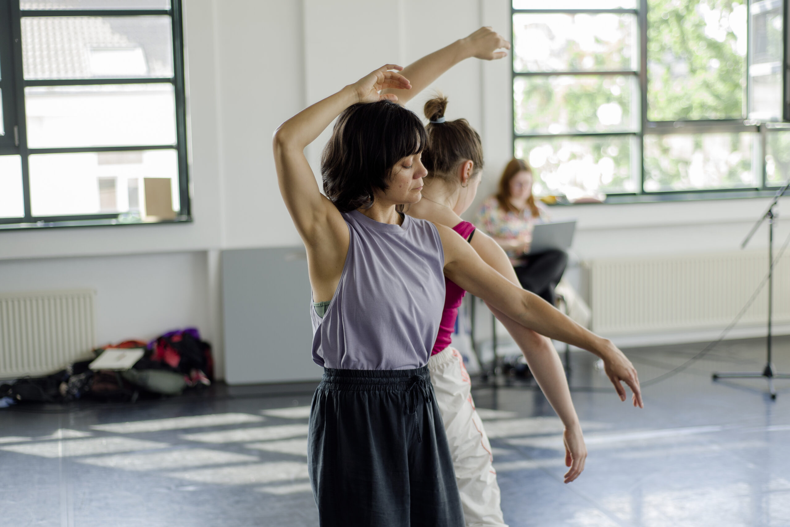Zwei Menschen tanzen Rücken an Rücken in einem hellen Raum. Sie tragen sportliche Kleidung. Im Hintergrund sitzt eine Person auf einem Stuhl mit Laptop auf dem Schoß.