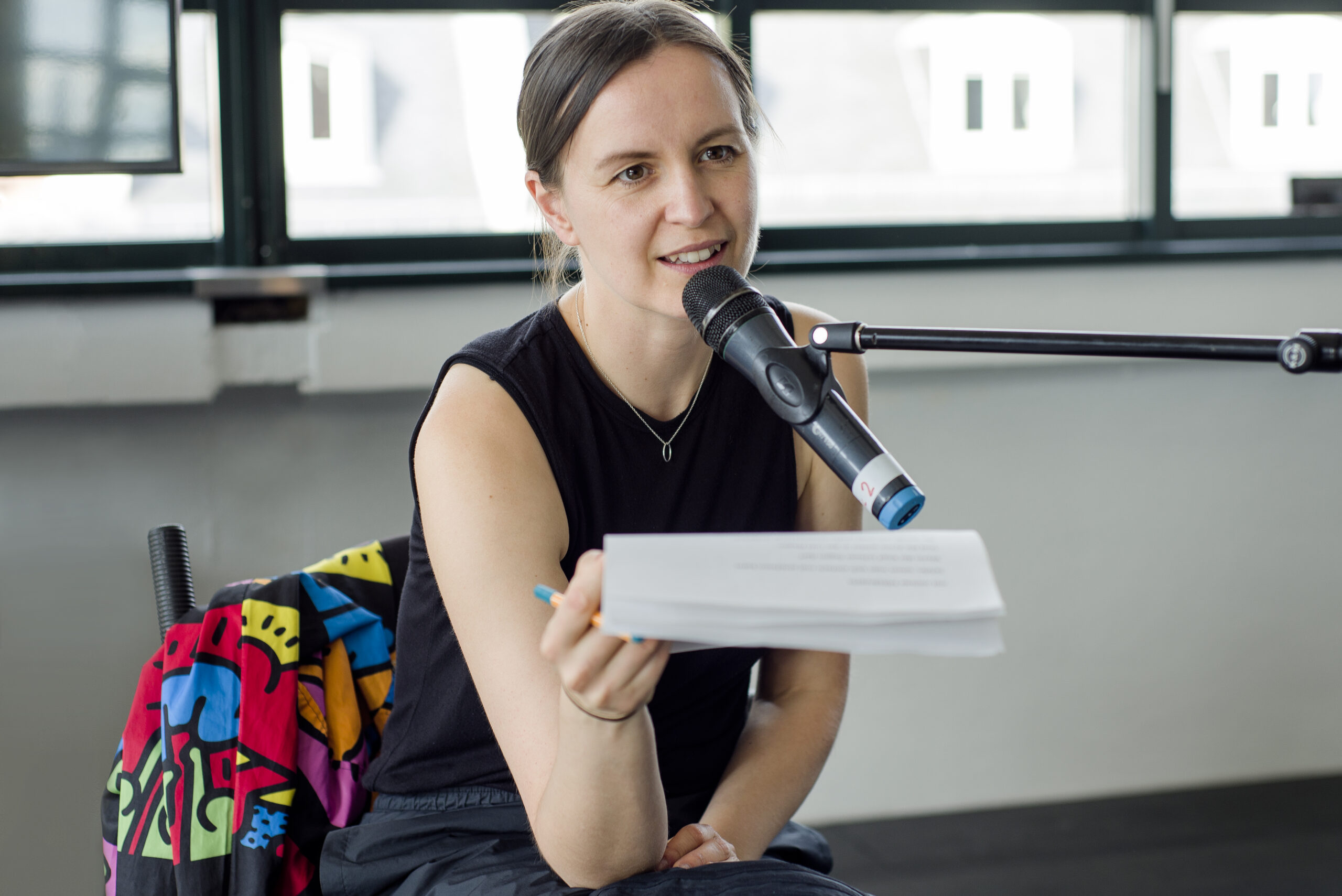 Eine Person spricht in ein Mikrofon. Sie sitzt und hält mehrere Blätter und einen Stift in der Hand.