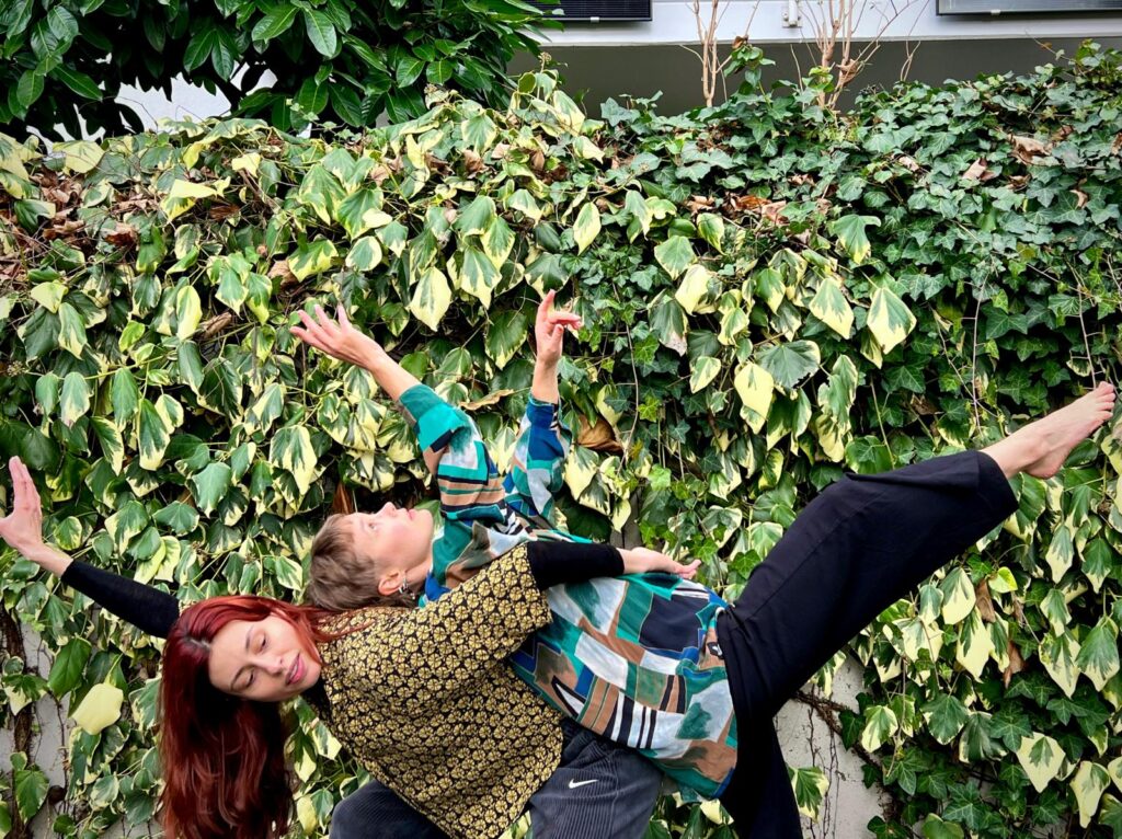 Zwei Personen vor einer mit Efeu bepflanzten Mauer. Sie tragen beide bunte Kleidung. Die eine streckt die Arme in die Luft und wird auf dem Rücken der anderen gehalten.