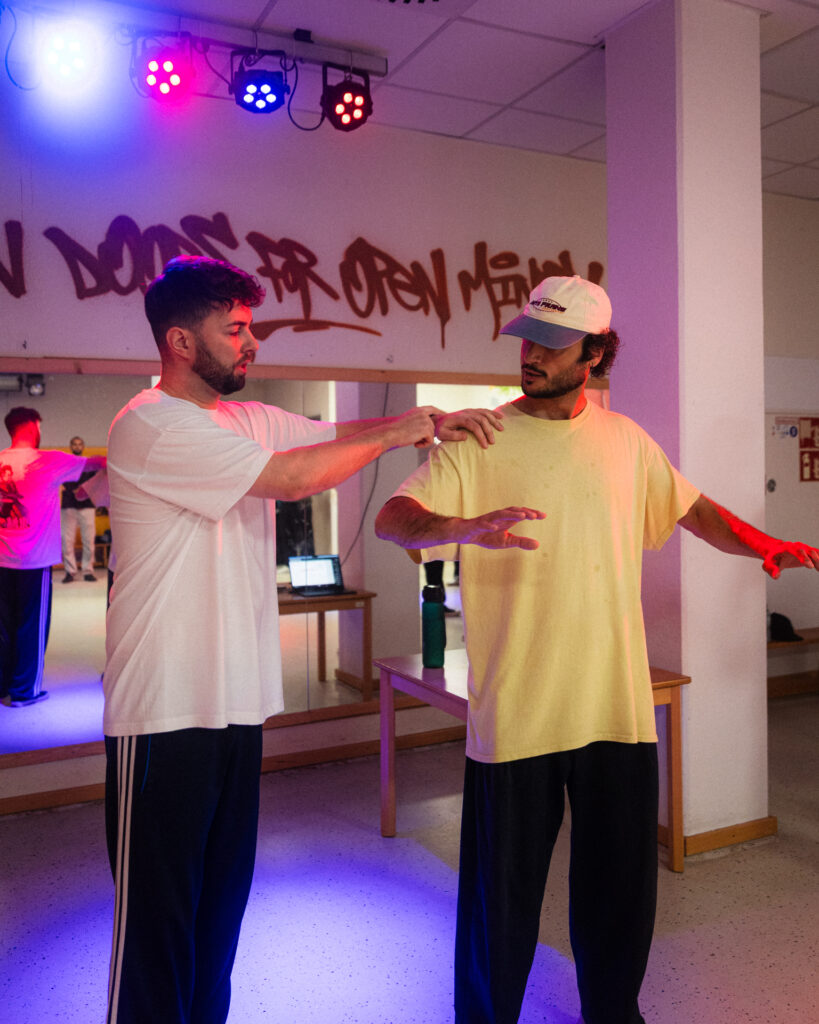 Zwei Männer in sportlicher Kleidung stehen in einem Tanzstudio. Der eine fasst dem anderen an die Schulter und zeigt wie er eine Bewegung ausführen soll.