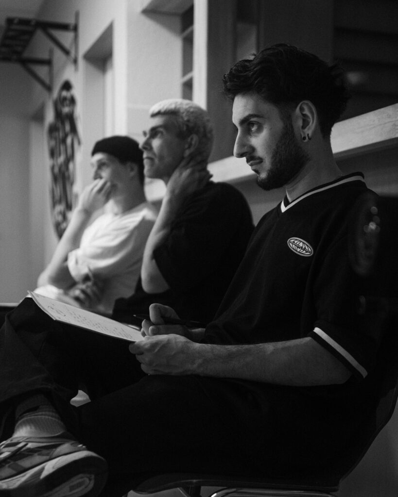 Ein Schwarzweiß Foto von drei jungen Männern, die nebeneinander sitzen und etwas vor ihnen betrachten.