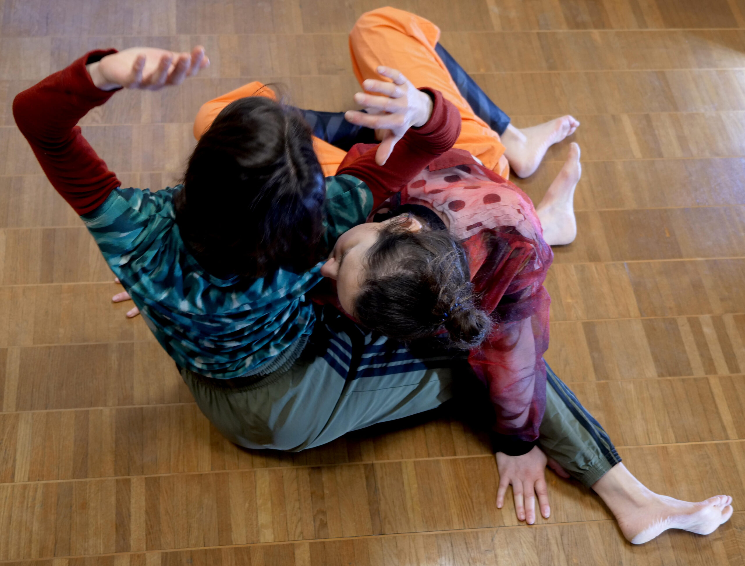 Das Bild zeigt zwei Personen, die sich akrobatisch übereinander auf einem Holzboden bewegen. Sie sind beide barfuß und tragen bunte Kleidung.
