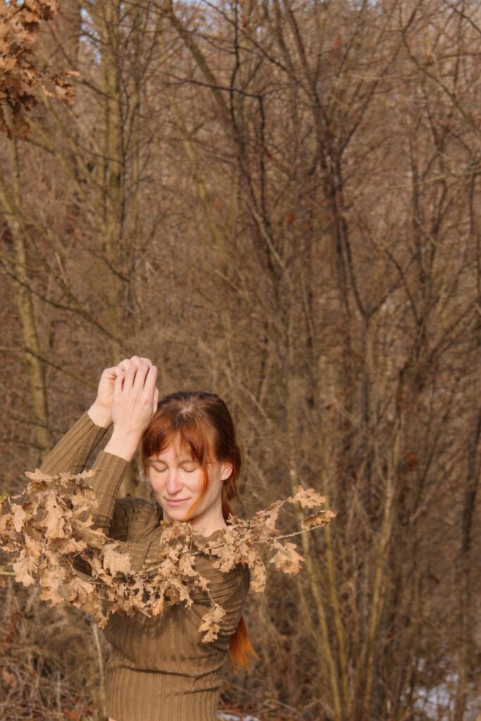 Ein Person tanz vor einem Waldhintergrund. Die Böume sind braun und ohne Blätter. Vor ihr hängt ein Ast mit braunen Blätterm