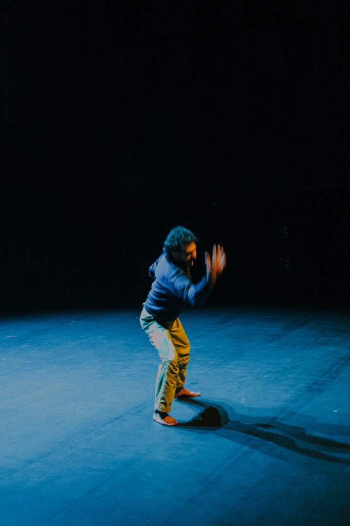 Auf dem Bild ist eine Person in einem Bühnenraum zu sehen. Das Licht ist bläulich. Die Person hat dunkle Haare und einen Bart, trägt ein blaues Shirt und eine beige Hose. Sie ist gerade in einer Bewegung und bewegt ihre Hand weg vom Kopf.