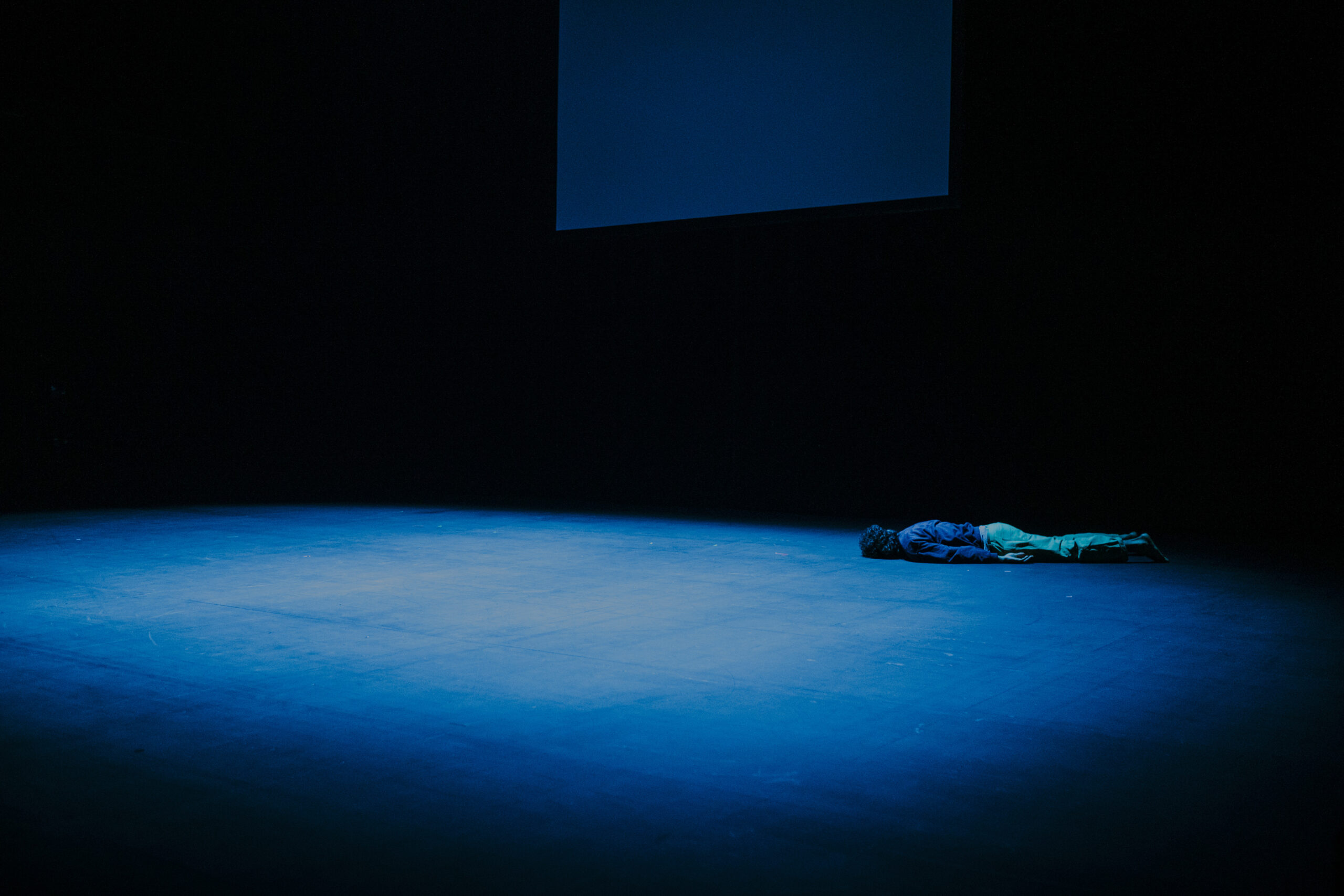 Eslam Enebishy liegt auf einer dunklen Bühne auf dem Bauch, mit dem Kopf zur hinteren Wand gedreht.